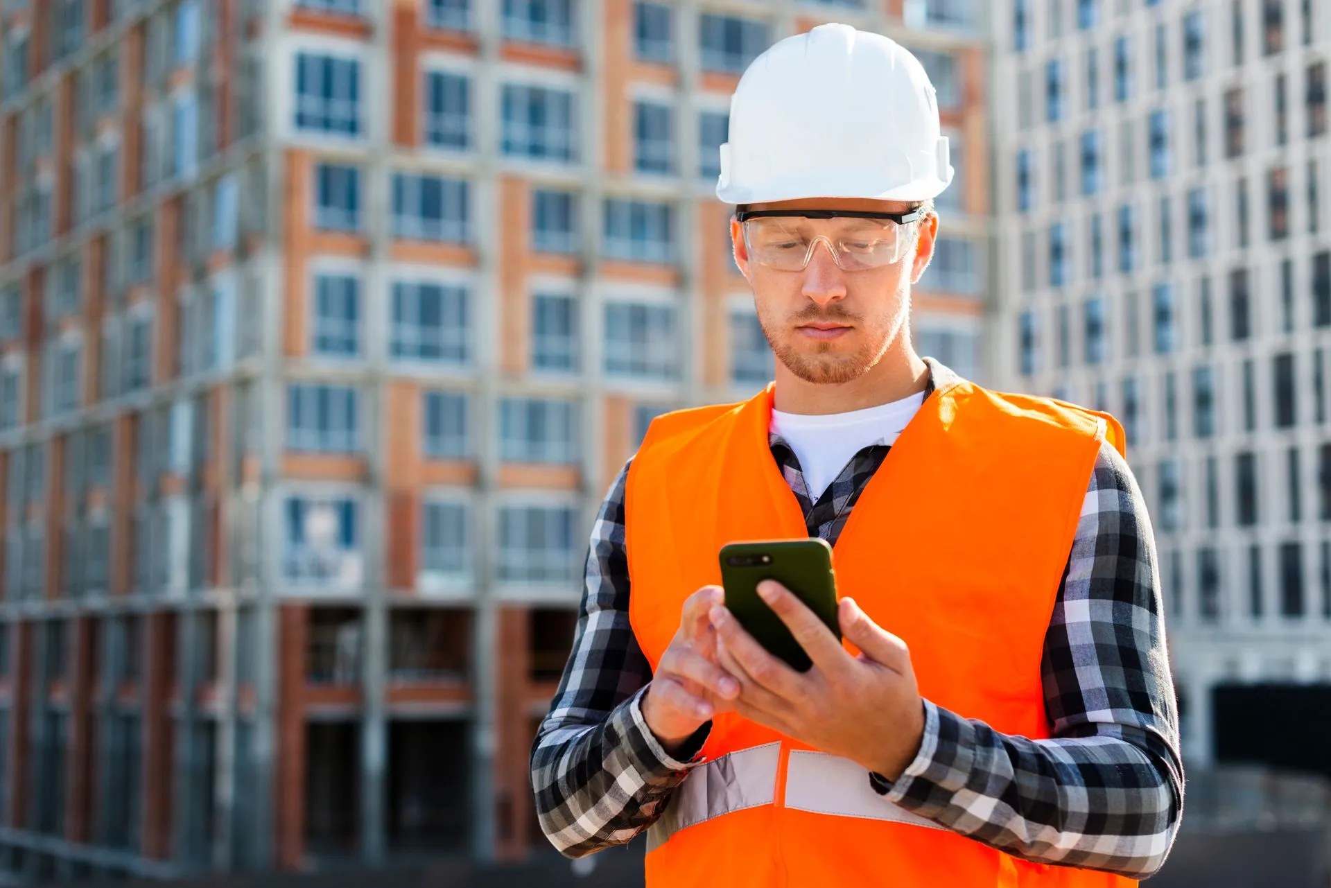 Un charpentier en casque blanc et veste orange qui consulte son téléphone sur un chantier, un immeuble en construction derrière lui